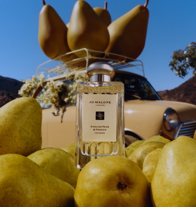 English Pear & Freesia cologne 100ml bottle surrounded by fresh pears and backdrop of large pear props on top of car rack.